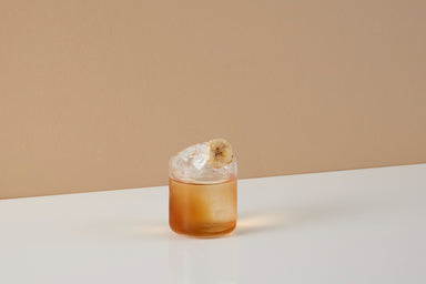 Image of a Craft Cocktails Banana Old Fashioned original recipe premixed ready to drink cocktail in a tumbler glass with ice and banana chip garnish shot on a brown and white background